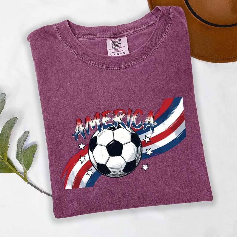 May include: A maroon short-sleeved t-shirt featuring a graphic of a soccer ball centered over red, white, and blue brushstroke stripes. The word AMERICA is printed in a distressed blue font above the ball, surrounded by small white stars.