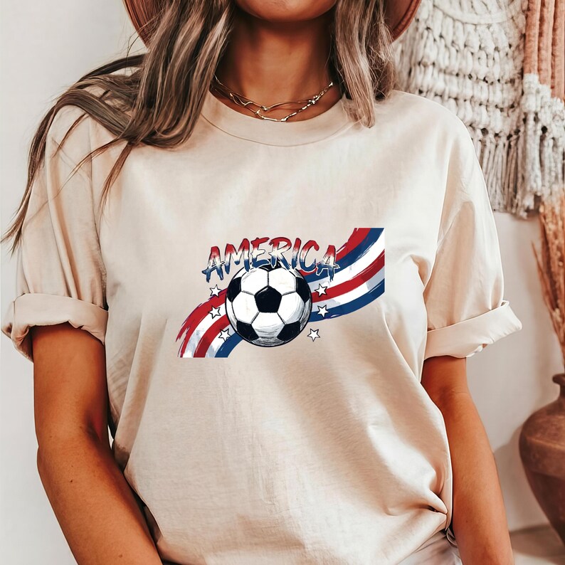 May include: A beige short-sleeve t-shirt featuring a graphic print of a soccer ball centered over red, white, and blue wavy stripes. The word AMERICA is printed in a distressed blue font above the ball, accented by small white stars.