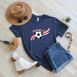 May include: A navy blue short-sleeve t-shirt featuring a graphic of a soccer ball centered over red, white, and blue wavy stripes with the word AMERICA printed above it, accompanied by small white stars scattered around the design.