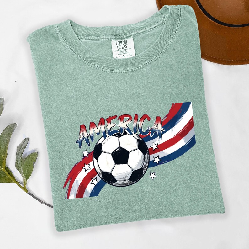 May include: A sage green short-sleeve t-shirt featuring a graphic of a black and white soccer ball centered over a red, white, and blue brushstroke design. The word AMERICA is printed in a distressed blue font above the soccer ball with small white stars.