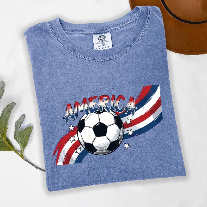 May include: A blue short-sleeved t-shirt featuring a graphic of a soccer ball centered over red, white, and blue wavy stripes and stars. The word AMERICA is printed in a distressed font above the ball. The shirt has a Comfort Colors brand label.