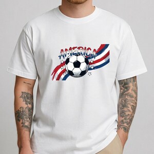 May include: A white short-sleeved t-shirt featuring a graphic of a soccer ball centered over red, white, and blue wavy stripes. The word AMERICA is printed in a stylized blue font above the ball, accompanied by small white star outlines.