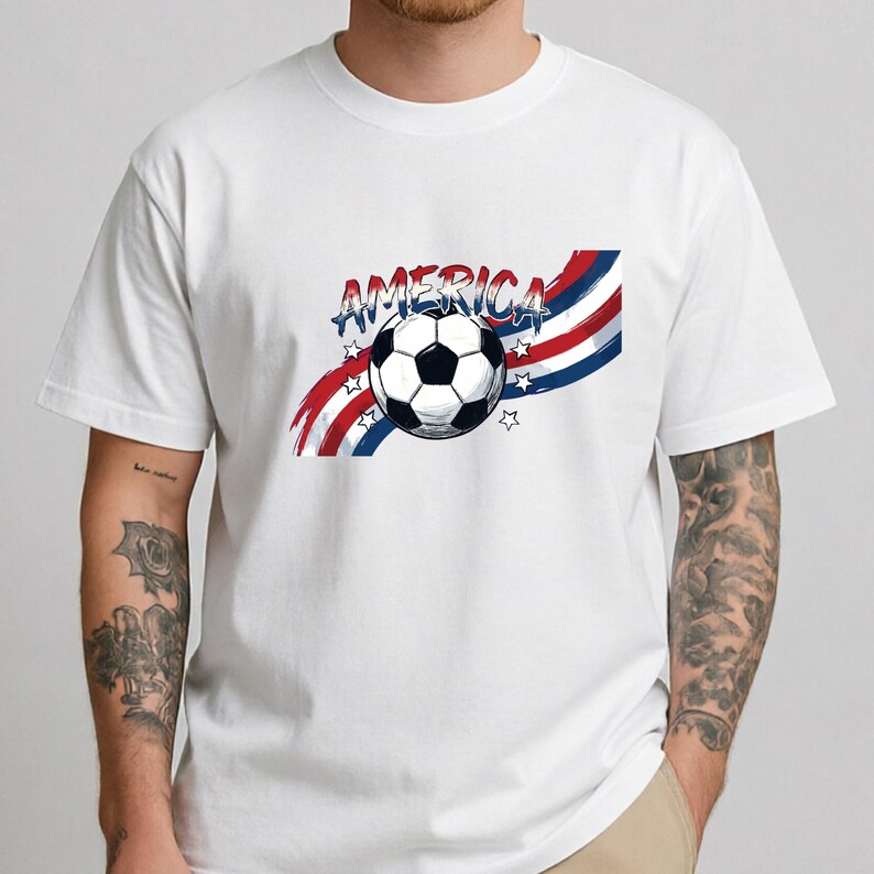 May include: A white short-sleeved t-shirt featuring a graphic of a soccer ball centered over red, white, and blue wavy stripes. The word AMERICA is printed in a stylized blue font above the ball, accompanied by small white star outlines.
