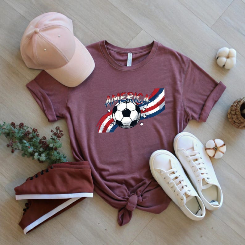 May include: A mauve short-sleeve t-shirt featuring a graphic of a soccer ball centered over red, white, and blue wavy stripes with the word AMERICA printed above. The shirt is displayed with a light pink baseball cap and white sneakers.