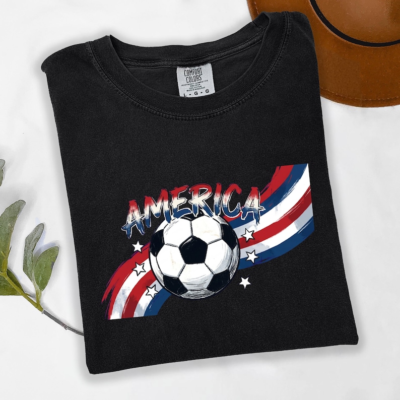 May include: A black short-sleeved t-shirt featuring a graphic of a soccer ball centered over a red, white, and blue brushstroke design. The word AMERICA is printed in a stylized font above the ball, accompanied by small white stars.