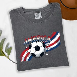 May include: A charcoal grey t-shirt featuring a graphic print of a black and white soccer ball centered over a red, white, and blue wavy striped background with the word AMERICA in a distressed blue font and several small white stars.