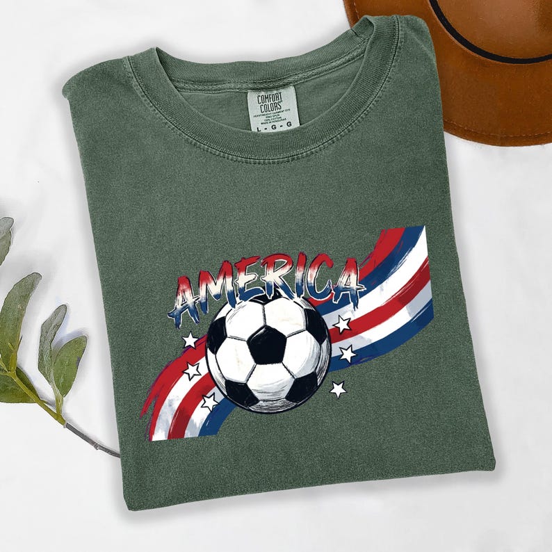 May include: A sage green t-shirt featuring a graphic of a black and white soccer ball centered over a red, white, and blue wavy striped background. The word AMERICA is printed in a distressed blue font above the ball, accented by small white stars.