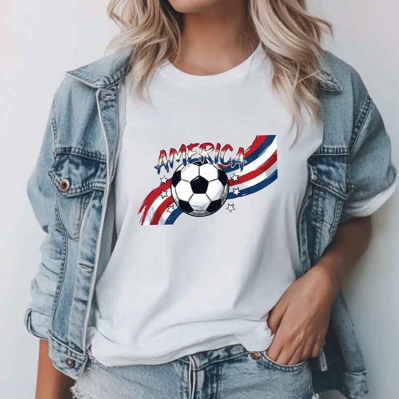 May include: A white t-shirt featuring a graphic of a black and white soccer ball centered over red, white, and blue wavy stripes. The word AMERICA is printed in a distressed blue font above the ball, accented by small white stars on the sides.