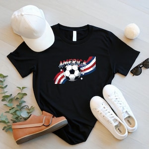 May include: A black t-shirt featuring a graphic of a soccer ball centered over red, white, and blue wavy stripes with the word AMERICA printed above the ball, accompanied by small white stars on a black background.