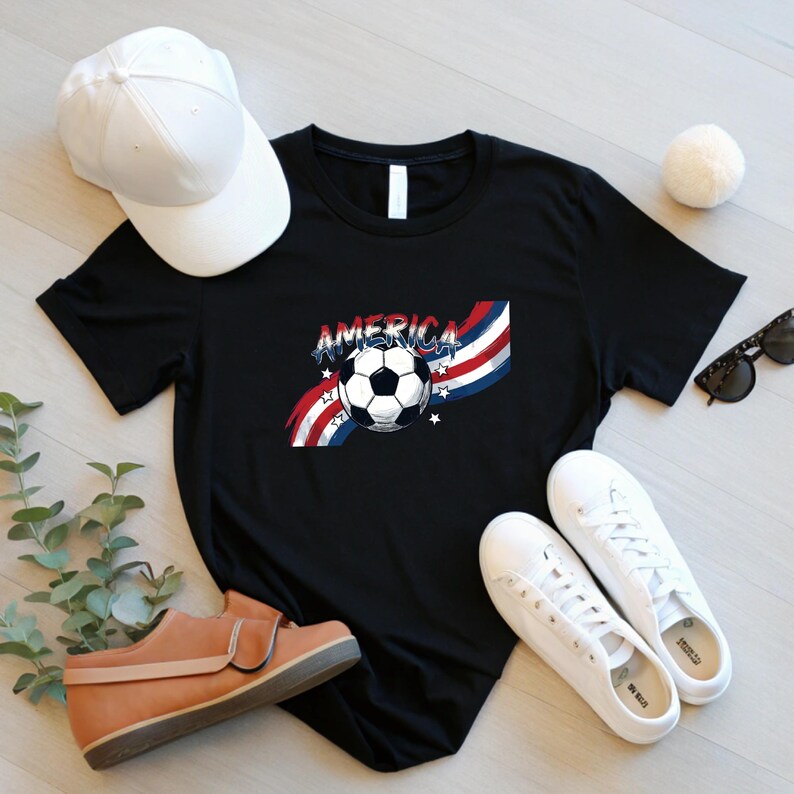 May include: A black t-shirt featuring a graphic of a soccer ball centered over red, white, and blue wavy stripes with the word AMERICA printed above the ball, accompanied by small white stars on a black background.