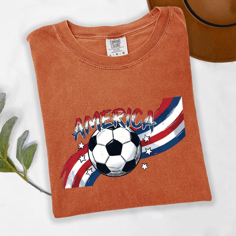 May include: A rust-colored t-shirt featuring a graphic of a soccer ball centered over a red, white, and blue brushstroke design with stars and the word AMERICA in a bold font. The shirt has a Comfort Colors label indicating a size large.