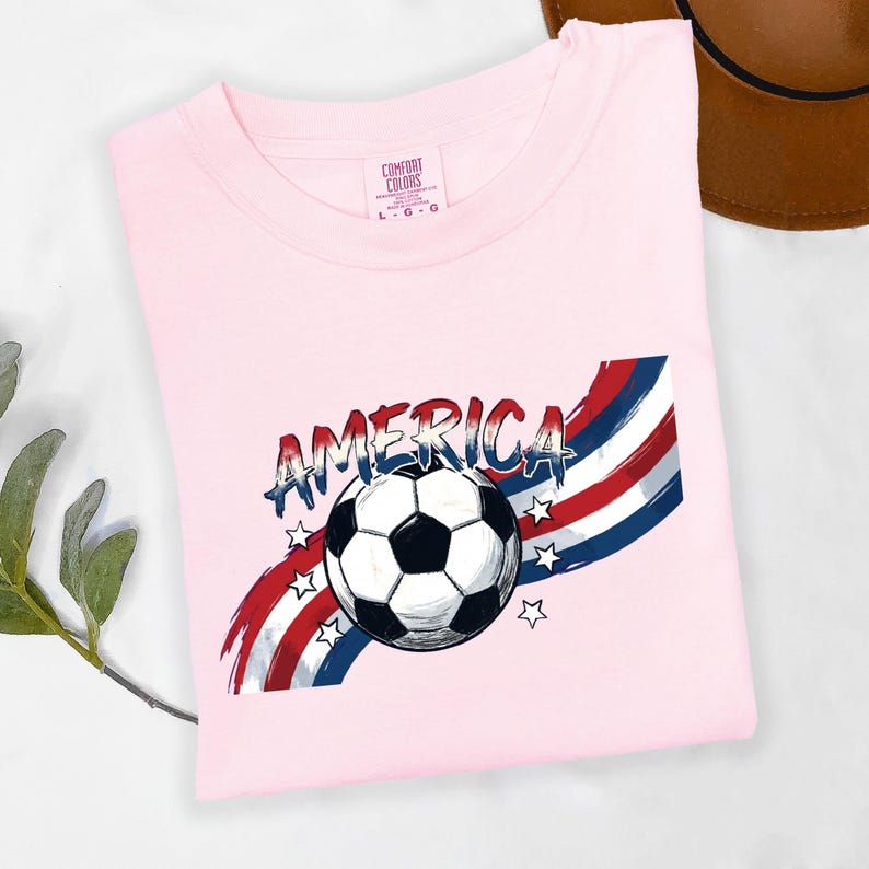 May include: A light pink t-shirt featuring a graphic of a black and white soccer ball centered over red, white, and blue brushstroke stripes. The word AMERICA is printed in a distressed blue font above the ball, accented by small white stars.