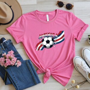 May include: A pink short-sleeved t-shirt featuring a graphic of a soccer ball centered over red, white, and blue wavy stripes. The word AMERICA is printed in a distressed blue font above the ball, accented by small white stars on both sides.
