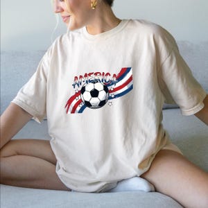May include: A cream-colored short-sleeve t-shirt featuring a graphic print of a black and white soccer ball centered over red, white, and blue wavy stripes and stars. The word AMERICA is printed in a distressed blue font above the soccer ball.