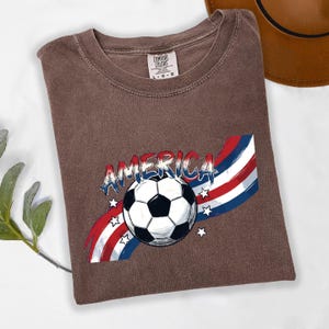 May include: A brown short-sleeved t-shirt with a graphic print featuring a black and white soccer ball centered over a red, white, and blue wavy striped background with stars and the word AMERICA written in a distressed blue font above the ball.