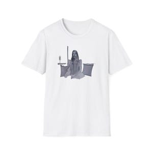 May include: White t-shirt with a grayscale illustration of a person seated with a long object and two pillows. A candle holder is also visible. The shirt has a classic crew neck and short sleeves.