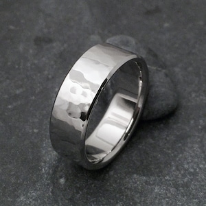 May include: A silver-toned metal ring featuring a wide band with a hammered, textured finish. The surface of the ring reflects light, highlighting the irregular, indented pattern across the exterior of the band, which has a smooth, polished interior.
