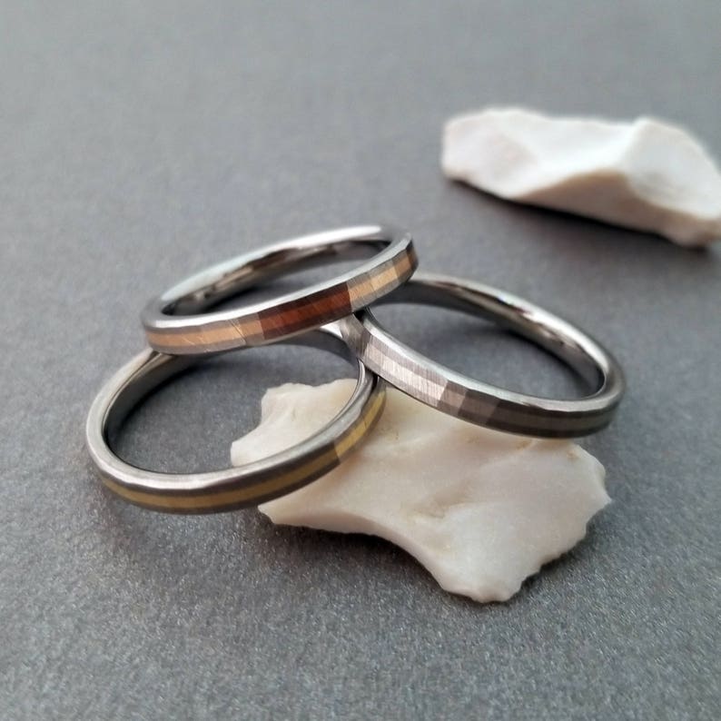 Puede incluir: Tres anillos de plata con finas bandas doradas y de cobre. Los anillos est&aacute;n apilados uno encima del otro y descansan sobre una roca blanca.