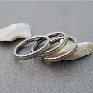 Puede incluir: Tres anillos de plata apilados uno encima del otro. El anillo superior tiene una fina banda dorada alrededor.
