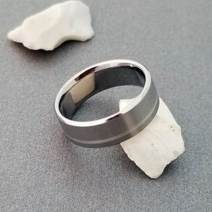 May include: A metallic ring with a brushed gray finish and polished beveled edges. The band features a subtle horizontal groove running around the center, creating a distinct two-tone effect with the reflective edges and the matte central surface.