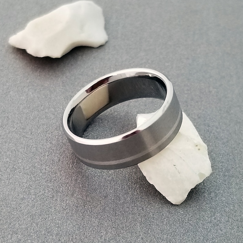 May include: A metallic ring with a brushed gray finish and polished beveled edges. The band features a subtle horizontal groove running around the center, creating a distinct two-tone effect with the reflective edges and the matte central surface.