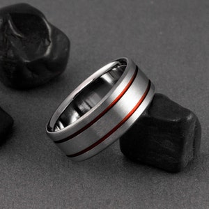 May include: A silver ring with two thin red lines around the band.