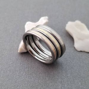Puede incluir: Tres anillos apilados, uno con una incrustaci&oacute;n de oro, sobre una superficie gris.