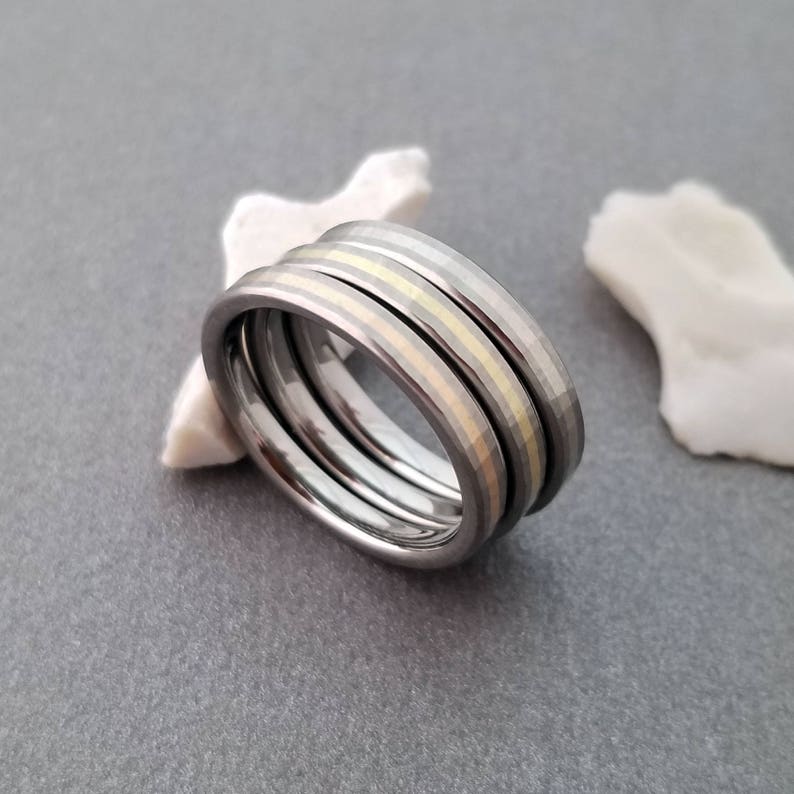 Puede incluir: Tres anillos apilados, uno con una incrustaci&oacute;n de oro, sobre una superficie gris.