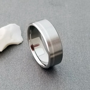 May include: A silver-toned metal ring with a wide band featuring a brushed matte finish on the exterior surface and a polished, reflective interior. The band has a distinct, thin, smooth groove running horizontally around the center of the ring.