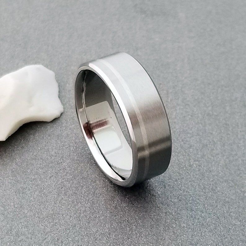May include: A silver-toned metal ring with a wide band featuring a brushed matte finish on the exterior surface and a polished, reflective interior. The band has a distinct, thin, smooth groove running horizontally around the center of the ring.