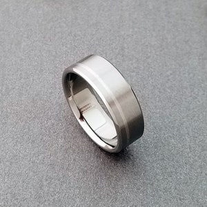 May include: A silver-toned metal ring featuring a wide, flat band with a brushed matte finish on the exterior surface and a polished, reflective interior. The edges of the band are beveled, creating a distinct border that catches the light.