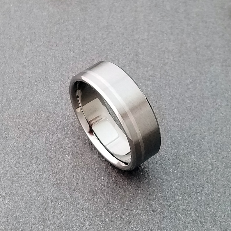 May include: A silver-toned metal ring featuring a wide, flat band with a brushed matte finish on the exterior surface and a polished, reflective interior. The edges of the band are beveled, creating a distinct border that catches the light.