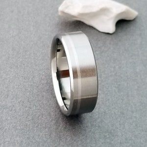 May include: A silver-toned metal ring with a flat profile and a brushed finish. The band features a single, thin, polished vertical groove running around the center, creating a subtle contrast against the matte surface of the metallic band.