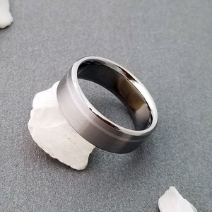 May include: A wide, circular metal ring with a brushed, matte gray finish on the outer band and a polished, reflective inner surface. The ring features a subtle, thin horizontal groove running along the center of the exterior band, creating a distinct visual separation.