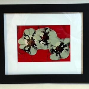 May include: Abstract floral art print in a black frame. The artwork features three stylized white flowers with black and brown centers against a vibrant red background. The print is matted in white.