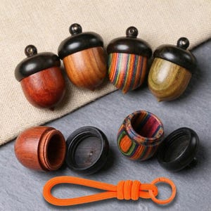 May include: A collection of wooden acorn-shaped containers with black tops and varying wood tones. One acorn is made of colorful, layered wood. An orange lanyard with a keyring is at the bottom.