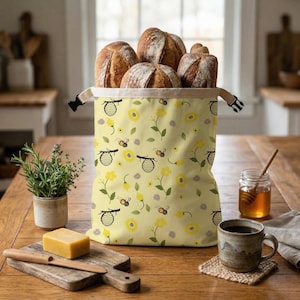 Puede incluir: Una bolsa de pan amarilla con un estampado de abejas y flores, llena de varias hogazas de pan. La bolsa está sobre una mesa de madera, junto a una pequeña planta, un tarro de miel y una taza de café.