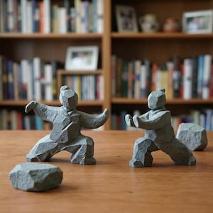 May include: Two grey stone sculptures of figures in martial arts poses, with two additional stone pieces. The figures are on a wooden surface, with a bookshelf in the background. The sculptures are detailed and textured.