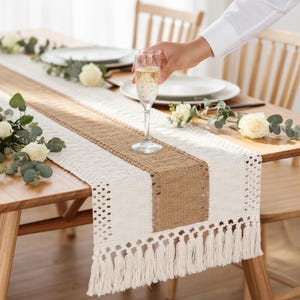 May include: A table runner with a central burlap stripe and white crochet sides. The runner has a fringe at the ends. A person is holding a champagne flute. The table is set with plates and flowers.
