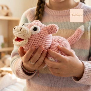 Op de afbeelding: Een handgemaakt roze gehaakt nijlpaard speelgoed met witte accenten, zwarte ogen en een rode mond. Het pluche dier wordt in de handen van een persoon gehouden. Het speelgoed is een zacht, knuffelbaar item, perfect voor kinderen.