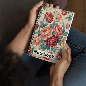 May include: A spiral-bound notebook with a floral pattern featuring red, pink, and peach roses with green leaves on a cream background. The word "Notebook" is printed on a white label on the cover.