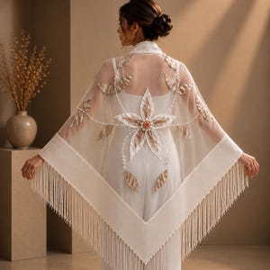May include: A sheer white shawl featuring a large central floral appliqué with gold-toned fabric petals, pearl accents, and a small peach-coloured centre. The triangular garment is finished with long, delicate fringe along the bottom edges.