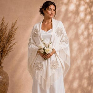 Peut inclure: Un châle de couleur crème orné de broderies circulaires complexes avec des accents centraux en forme de perles et une longue frange sur les bords. Le châle est drapé sur une robe sans manches de couleur crème, créant un aspect superposé.