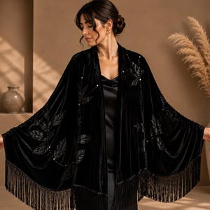 Puede incluir: Un chal de terciopelo negro con frente abierto, flecos largos en el dobladillo inferior y bordados en forma de hoja en tono sobre tono acentuados con pequeñas cuentas, usado sobre un vestido lencero de satén negro con cuello desbocado.