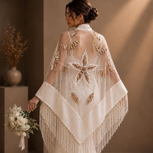 Puede incluir: Un chal blanco transparente con bordados dorados en forma de hoja y un motivo floral central con detalles de perlas. La prenda triangular tiene largos flecos en los bordes y se lleva sobre un vestido largo de encaje hasta el suelo.