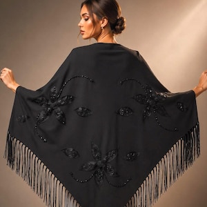 May include: A black triangular shawl featuring three large floral appliqués made of dark beads and sequins. The edges of the garment are finished with long, thin black fringe that hangs down from the bottom point and sides of the fabric.
