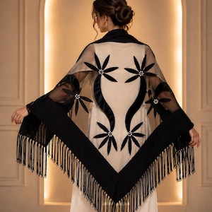 May include: A black sheer shawl featuring velvet floral appliqués with pearl centers and a wide black border with long fringe. The shawl is draped over a plain white garment, showcasing a symmetrical design of black velvet petals and stems.
