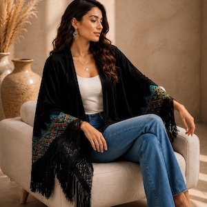 A black velvet shawl with colorful embroidered patterns on the sleeves and fringe trim. The shawl is worn over a white top and blue jeans. The sleeves have a geometric design in shades of teal, orange, and gold.