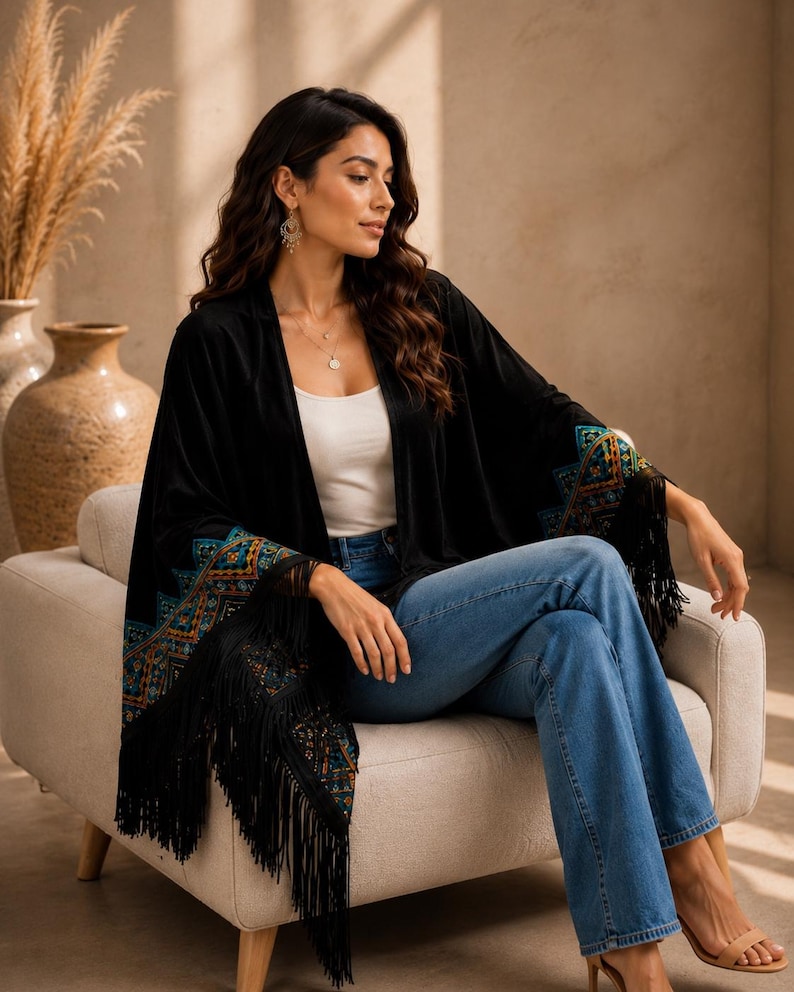 A black velvet shawl with colorful embroidered patterns on the sleeves and fringe trim. The shawl is worn over a white top and blue jeans. The sleeves have a geometric design in shades of teal, orange, and gold.