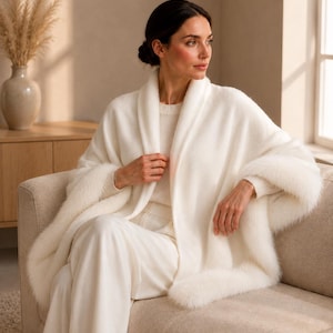 A cream-colored open-front shawl featuring a soft, fuzzy texture and a wide shawl collar. The garment is draped over the shoulders and arms, showcasing a plush, fur-like trim along the edges and a smooth, knit-like body construction.